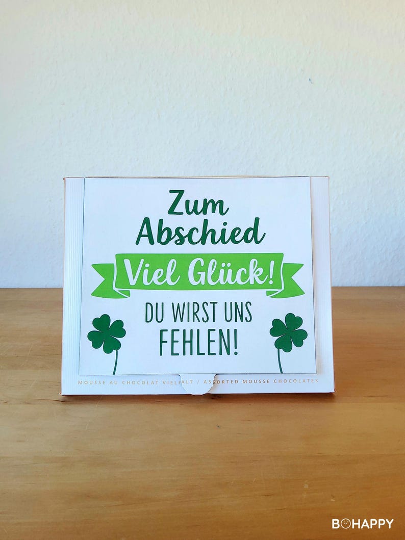 May include: A white box with a green and white label. The label has the text "Zum Abschied Viel Gl&uuml;ck! DU WIRST UNS FEHLEN!" and two green shamrock illustrations. The box is on a wooden surface.