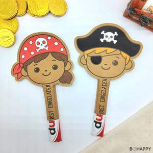 Könnte beinhalten: Zwei Piraten-Papierausstanzungen. Eines zeigt ein Piratenmädchen mit rotem Bandana und Totenkopf. Das andere ist ein Piratenjunge mit schwarzem Hut und Augenklappe. Beide haben den Text "DU BIST EINGELADEN" auf dem Stiel.
