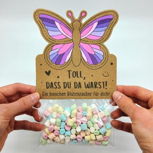 Può includere: Un'etichetta regalo a forma di farfalla con un sacchetto di marshmallow pastello. L'etichetta è marrone con un disegno di farfalla viola, rosa e blu. Il testo sull'etichetta recita "Toll, dass du da warst! Ein bisschen Blutenzauber für dich!"