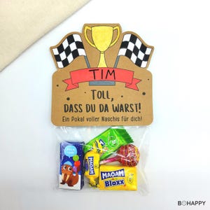 May include: A clear cellophane bag filled with candy, attached to a brown cardboard sign. The sign features a gold trophy, checkered flags, and the text "TIM, TOLL, DASS DU DA WARST! Ein Pokal voller Naschis für dich!" in red and black.