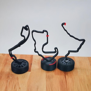 Op de afbeelding: Drie zwarte 3D-geprinte racebaanmodellen op zwarte bases met rode accenten. De banen zijn gevormd als verschillende racebanen en worden weergegeven op een houten oppervlak.