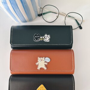 Op de afbeelding: Drie zwarte leren brillenetuis met verschillende geborduurde ontwerpen. De bovenste etui heeft een zwarte kat en een witte kat met een bierpul. De middelste etui heeft een bruine beer met een spraakballon. De onderste etui heeft een geel huis.