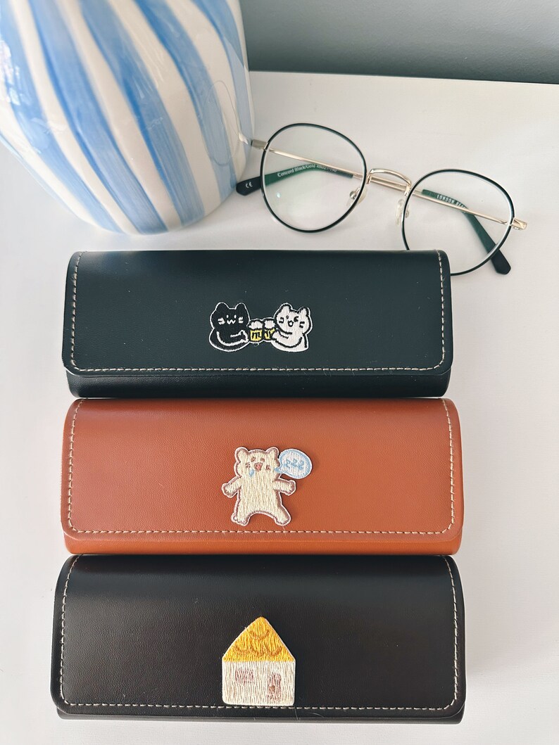 Op de afbeelding: Drie zwarte leren brillenetuis met verschillende geborduurde ontwerpen. De bovenste etui heeft een zwarte kat en een witte kat met een bierpul. De middelste etui heeft een bruine beer met een spraakballon. De onderste etui heeft een geel huis.