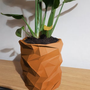 May include: A green plant with large leaves in a geometric orange planter. The planter is made of a textured material and has a unique, modern design.