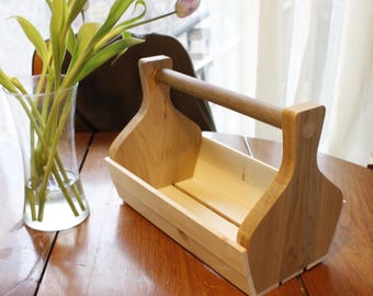 May include: A wooden tool caddy with a handle. The caddy is made of light brown wood and has a rectangular shape. It has a divider in the middle to separate items.