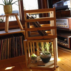 May include: Wooden crates stacked to display vinyl records and potted plants. The crates are made of light-colored wood and are arranged in a room with natural light. A small wooden stool holds a potted plant. The room has a vintage aesthetic.