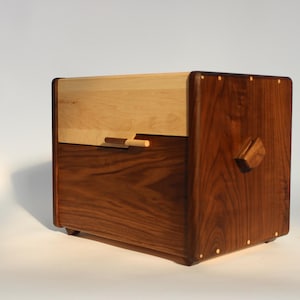 May include: A handcrafted wooden box with a light-colored top and a darker, walnut-toned base. The box features a small, rectangular handle and a decorative wooden accent on the side. The corners are rounded, and the box sits on small feet.