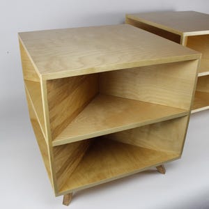 May include: Two light-colored wooden nightstands with a modern design. Each nightstand has a rectangular top, two shelves, and angled sides. The nightstands are supported by small, angled wooden legs. The wood grain is visible.