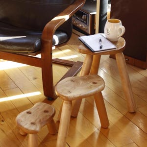 May include: Three small, handcrafted wooden stools with rounded seats and three legs. One stool has a notepad, pen, and a mug on top. The stools are made of light-colored wood and are placed on a hardwood floor.