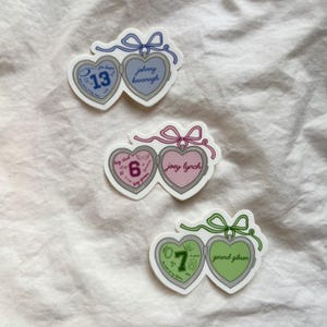 May include: Three heart-shaped stickers with a locket design. Each sticker has a different color scheme: blue and gray, pink and light pink, and green and light green. Each locket has a number and a name written on it.