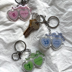 May include: Four heart-shaped keyrings with silver rings. Each keyring has a different colour scheme: pink, blue, and green. The keyrings have text that includes names and numbers. The keyrings are on a white surface.