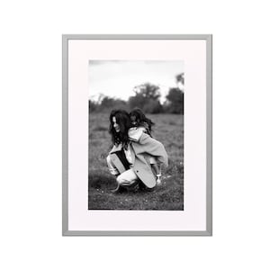 Könnte beinhalten: Schwarz-weiß gerahmtes Foto einer Frau, die ein Kind auf dem Rücken in einem Feld trägt. Die Frau trägt einen Mantel und eine Hose. Das Kind trägt einen Mantel und Schuhe. Das Foto ist in einem silbernen Rahmen.