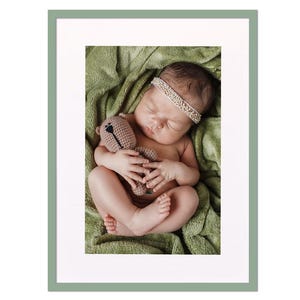 Op de afbeelding: Een pasgeboren baby slaapt op een groene deken en houdt een bruine gehaakte teddybeer vast. De baby draagt een wit parelbandje.