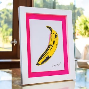 May include: A framed print of a yellow banana with black markings on a white background. The frame has a pink border and the print is signed "Andy Warhol".