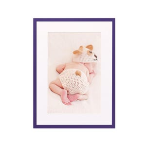 May include: A sleeping baby wearing a crocheted dog costume with a brown nose and ears. The baby is lying on a white blanket.