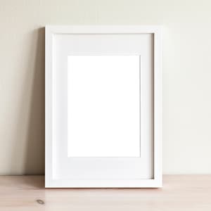 May include: A white picture frame with a white mat on a light wood surface.