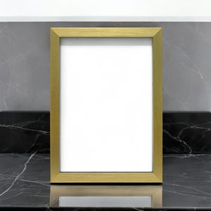 Può includere: Una cornice rettangolare dorata con interno bianco, posta su uno sfondo di marmo grigio e nero. La cornice è posizionata su una superficie di marmo nero, creando un contrasto di trame e colori.