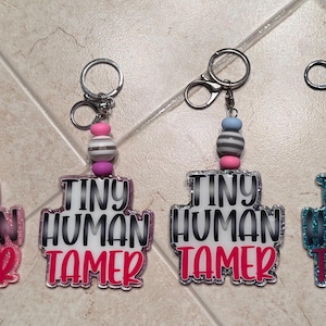 May include: Four keychains with the text "TINY HUMAN TAMER" in a bold font. Each keychain has a silver ring and a beaded accent in various colors, including pink, white, and blue. The keychains are on a tiled surface.