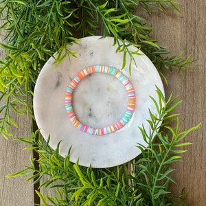 Könnte beinhalten: Ein farbenfrohes Armband aus kleinen, runden Perlen in den Farben Rosa, Orange und Blau. Das Armband liegt auf einer weißen Marmorfläche.