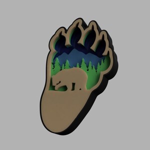 May include: A brown bear paw print with a black outline. Inside the paw print is a green forest scene with a brown bear walking through it. The forest scene has blue mountains in the background.