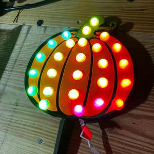 Op de afbeelding: Een houten bord in de vorm van een pompoen met een zwarte omtrek en een groene steel. De pompoen is versierd met 20 kleurrijke LED-lampjes, waaronder rood, oranje, geel, groen, blauw en paars.