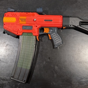 May include: A red and black toy blaster with an orange trigger and accents. The blaster has a large magazine and a black stock. The text "Adventure Force" is visible on the side of the blaster.