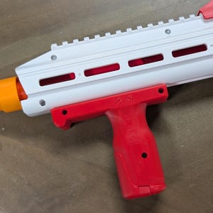 May include: A white toy blaster featuring a bright red vertical foregrip attached to the underside of the barrel. The barrel assembly includes a top rail, horizontal ventilation slots, and a vibrant orange tip at the front end of the toy weapon.