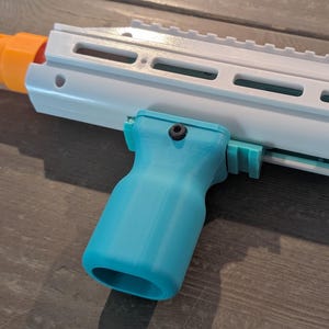 May include: A white toy blaster barrel featuring a bright orange muzzle tip and a teal vertical foregrip attached to the underside. The foregrip is secured with a single black screw and features a smooth, contoured shape for a firm grip.