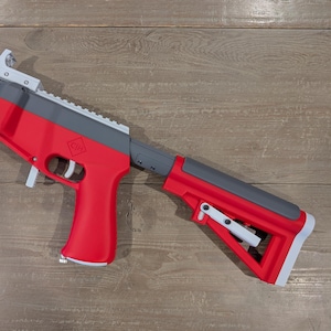 May include: A toy blaster featuring a red, gray, white, and orange color scheme. The device includes a red pistol grip, a red and gray adjustable shoulder stock, a gray top rail, and a front orange muzzle section with a vertical red handle.