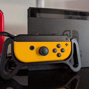 Peut inclure: Une manette Joy-Con Nintendo Switch jaune et noire avec une poign&eacute;e noire. Une manette Joy-Con rouge est visible &agrave; gauche. La console Nintendo Switch est &agrave; l'arri&egrave;re-plan.