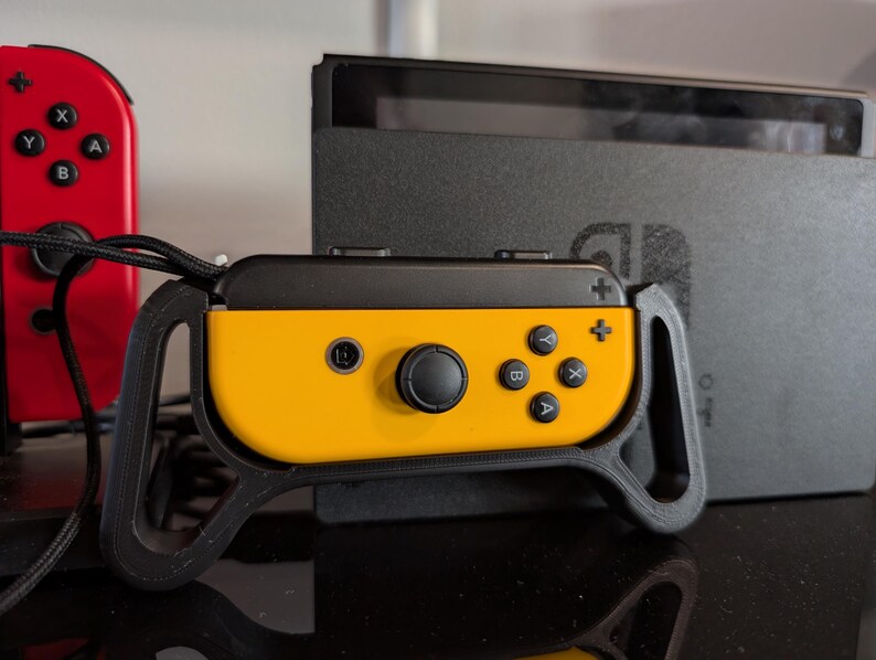 Peut inclure: Une manette Joy-Con Nintendo Switch jaune et noire avec une poign&eacute;e noire. Une manette Joy-Con rouge est visible &agrave; gauche. La console Nintendo Switch est &agrave; l'arri&egrave;re-plan.