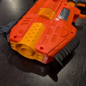May include: Orange and yellow toy gun with a black handle and accents. The toy gun has the word "ADVENTURE" printed on the side. The gun is made of plastic and has a textured surface.