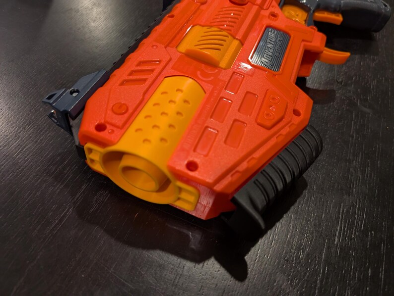 May include: Orange and yellow toy gun with a black handle and accents. The toy gun has the word "ADVENTURE" printed on the side. The gun is made of plastic and has a textured surface.