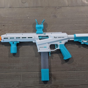 May include: A white and teal toy blaster with an orange muzzle tip. The blaster features a vertical foregrip, a top-mounted sight, a detachable magazine, and a stock. The side of the blaster is printed with the text GAME FACE and TRION.