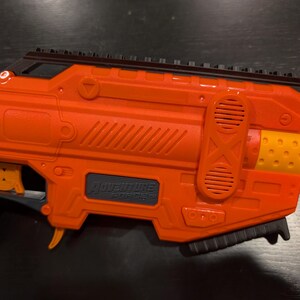 May include: An orange and black toy gun with the text "ADVENTURE FORCE" and "SPECTRUM" visible. The toy gun has a black top rail, a trigger, and a grip. The barrel end is yellow with small holes.