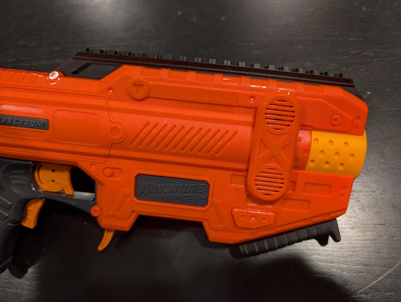 May include: An orange and black toy gun with the text "ADVENTURE FORCE" and "SPECTRUM" visible. The toy gun has a black top rail, a trigger, and a grip. The barrel end is yellow with small holes.