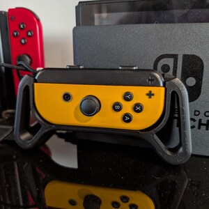Peut inclure: Manette Joy-Con Nintendo Switch jaune et noire avec une poign&eacute;e noire. La manette comporte des boutons noirs et un joystick noir. Une manette Joy-Con rouge est visible en arri&egrave;re-plan, ainsi qu'une console Nintendo Switch noire.