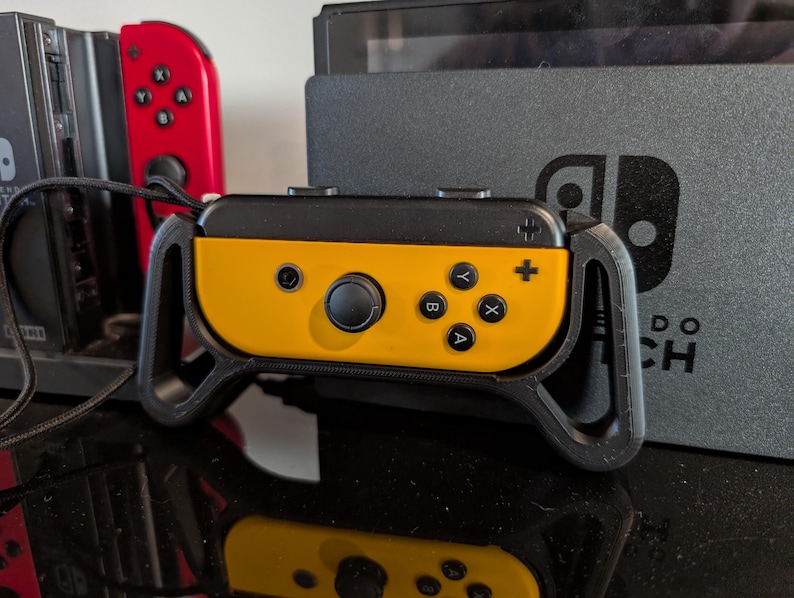 Peut inclure: Manette Joy-Con Nintendo Switch jaune et noire avec une poign&eacute;e noire. La manette comporte des boutons noirs et un joystick noir. Une manette Joy-Con rouge est visible en arri&egrave;re-plan, ainsi qu'une console Nintendo Switch noire.