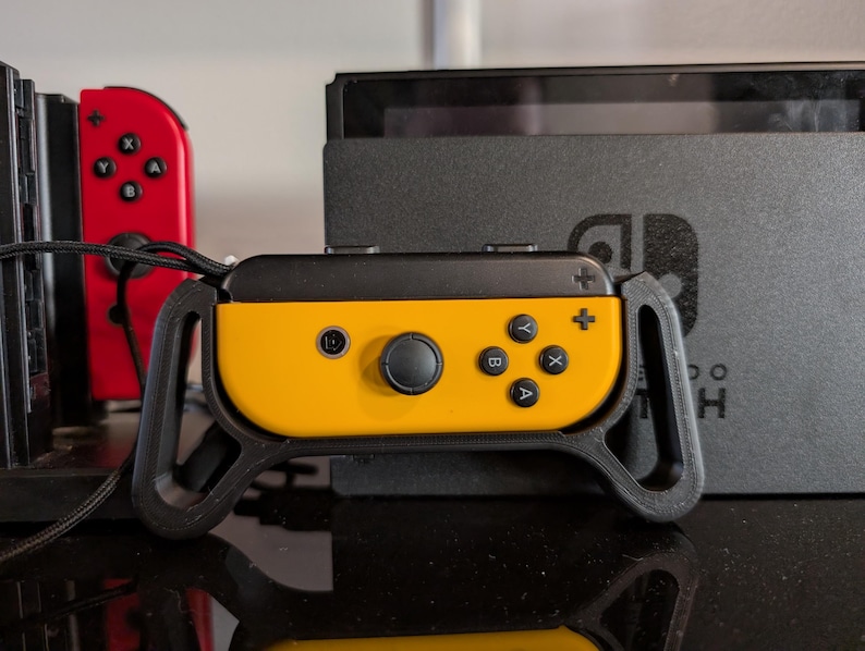 Peut inclure: Une poign&eacute;e de manette de jeu jaune et noire avec des boutons noirs et un joystick noir. Une manette de jeu rouge est visible &agrave; gauche. Une console de jeu noire est en arri&egrave;re-plan.