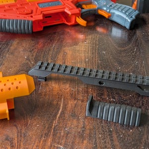 May include: An orange and red toy gun with gray and orange accents, along with black accessories. The accessories include a black rail and a black grip. The toy gun is on a wooden surface.