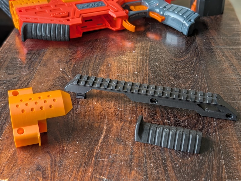 May include: An orange and red toy gun with gray and orange accents, along with black accessories. The accessories include a black rail and a black grip. The toy gun is on a wooden surface.