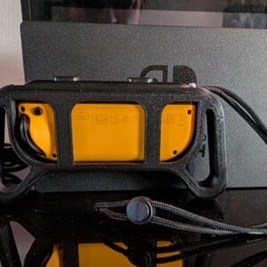 Peut inclure: Une poign&eacute;e de contr&ocirc;leur noire avec des accents jaunes, con&ccedil;ue pour une console de jeu. La poign&eacute;e pr&eacute;sente une surface textur&eacute;e et une lani&egrave;re pour une manipulation s&ucirc;re. La console est visible en arri&egrave;re-plan.