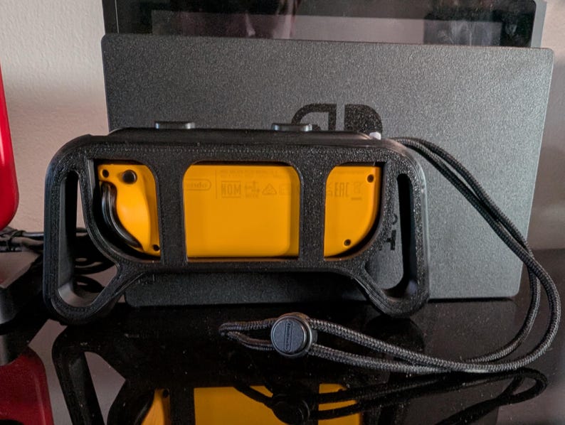 Peut inclure: Une poign&eacute;e de contr&ocirc;leur noire avec des accents jaunes, con&ccedil;ue pour une console de jeu. La poign&eacute;e pr&eacute;sente une surface textur&eacute;e et une lani&egrave;re pour une manipulation s&ucirc;re. La console est visible en arri&egrave;re-plan.