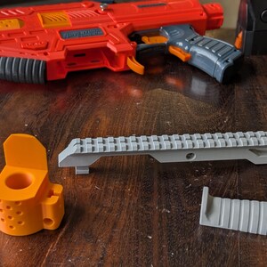 May include: An orange and gray toy blaster with accessories, including a gray rail mount, an orange mounting bracket, and a gray grip. The blaster is red and orange with a gray handle and black accents. The accessories are on a wooden surface.