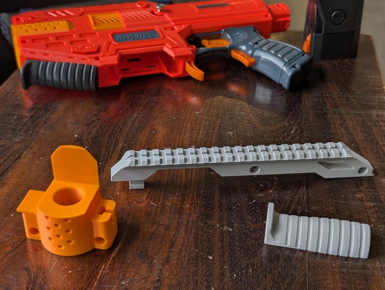 May include: An orange and gray toy blaster with accessories, including a gray rail mount, an orange mounting bracket, and a gray grip. The blaster is red and orange with a gray handle and black accents. The accessories are on a wooden surface.