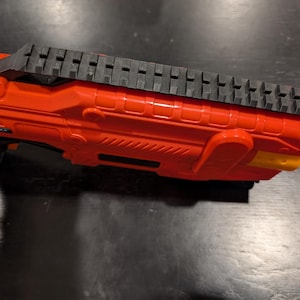 May include: Close-up of a bright orange toy gun with a black top rail. The gun has a textured design and a yellow accent on the end. The toy gun is made of plastic.