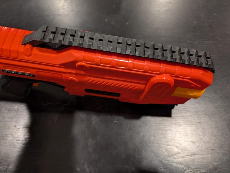May include: Close-up of a bright orange toy gun with a black top rail. The gun has a textured design and a yellow accent on the end. The toy gun is made of plastic.