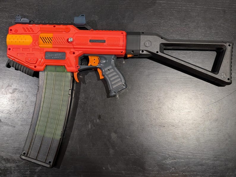 May include: A red and black toy blaster with an orange trigger and accents. The blaster has a large magazine and a black stock. The text "Adventure Force" is visible on the side of the blaster.