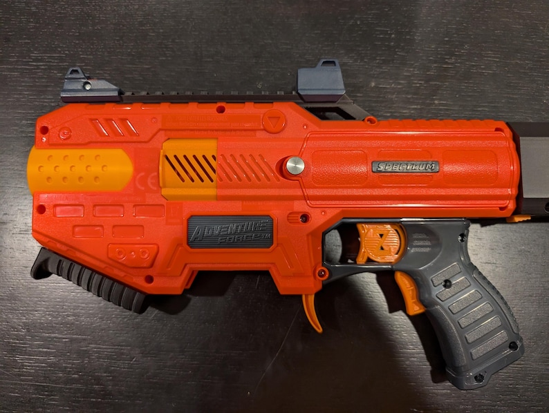 May include: Orange and gray Adventure Force Spectrum toy blaster. The toy gun has a black handle, orange trigger, and orange and yellow accents. The text "SPECTRUM" and "ADVENTURE FORCE" are visible on the blaster.