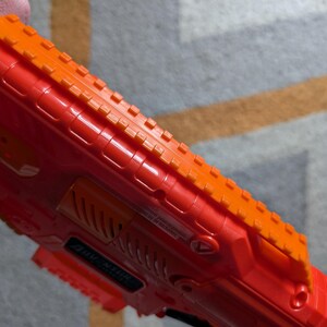May include: Close-up of a red and orange toy gun. The toy gun has a textured orange top rail and a red body. The gun has a series of ridges and grooves along its surface. The toy gun is held by a person.